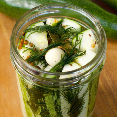 An open jar of zucchini pickle spears with dill, garlic, and spices on a table.